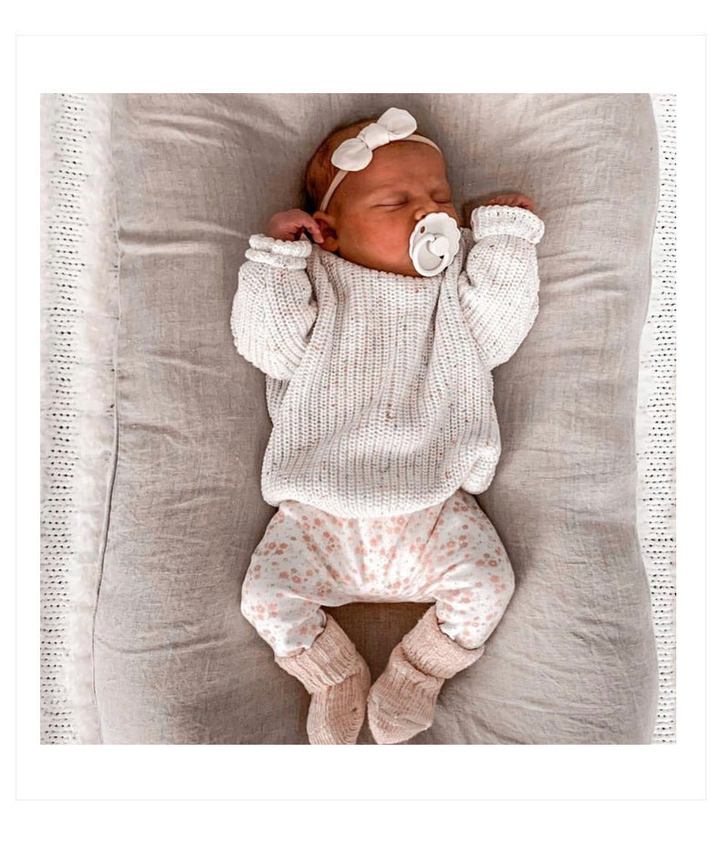 Newborn baby in a white knitted outfit with a pacifier and headband, lying on a gray blanket.