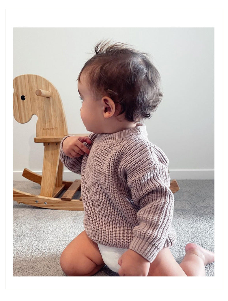 Baby wearing a knitted sweater sitting on the floor with a wooden rocking horse in the background.