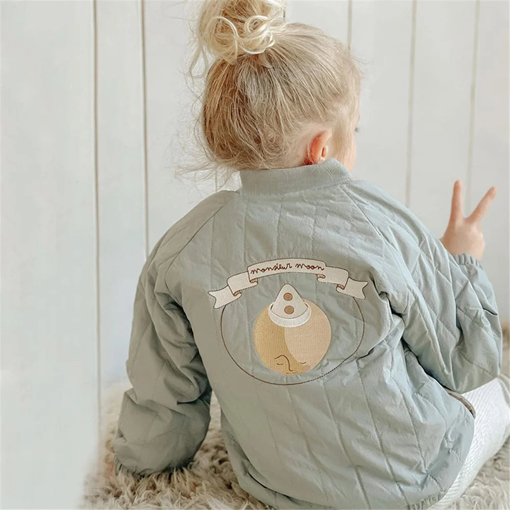 Child wearing a light blue jacket with a bear design and text on the back, sitting on a white floor.