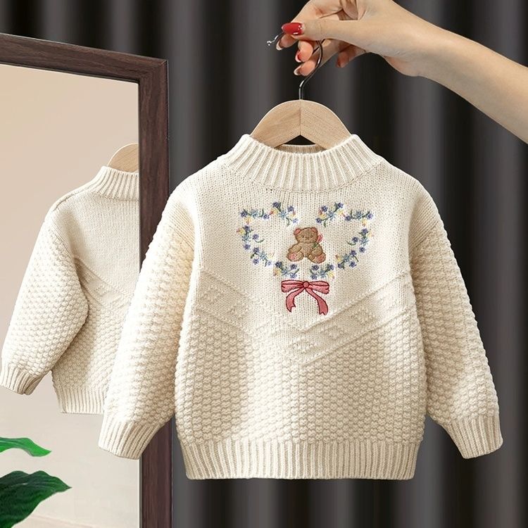 Children's sweater with teddy bear and floral design on a hanger against a dark curtain background.