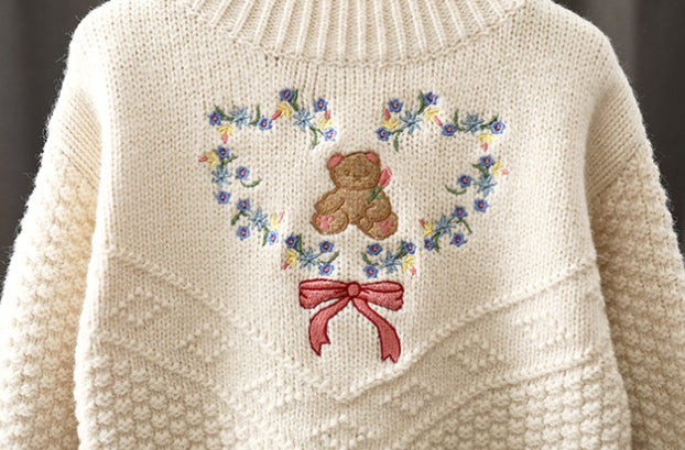 Cream-colored sweater with embroidered teddy bear and floral design on a dark background